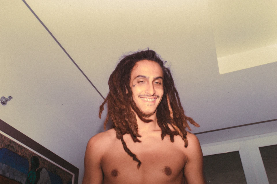 The image shows a young man with dreadlocks standing in front of a window, smiling at the camera. He is shirtless and has his hair styled in long dreadlocks that fall over his shoulders. The background features a white ceiling with a skylight, allowing natural light to illuminate the room. A painting can be seen hanging on the wall above the man's head.