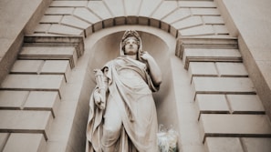 A classical stone statue of a woman draped in flowing robes, positioned in a niche of a stone building. The surrounding architecture features a symmetrical pattern of rectangular blocks, with an arched design above the statue. The statue holds a scroll and has an expression of serene contemplation. At the base of the statue, there is a bouquet of flowers.