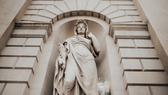 A classical stone statue of a woman draped in flowing robes, positioned in a niche of a stone building. The surrounding architecture features a symmetrical pattern of rectangular blocks, with an arched design above the statue. The statue holds a scroll and has an expression of serene contemplation. At the base of the statue, there is a bouquet of flowers.