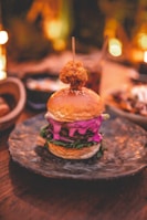 A gourmet burger is presented on a textured, dark plate. The burger features a sesame seed bun with vibrant, colorful fillings, including pink sauce and green leafy vegetables. A skewer holds a crispy fried element on top. The setting is warmly lit, suggesting an intimate or cozy dining environment.