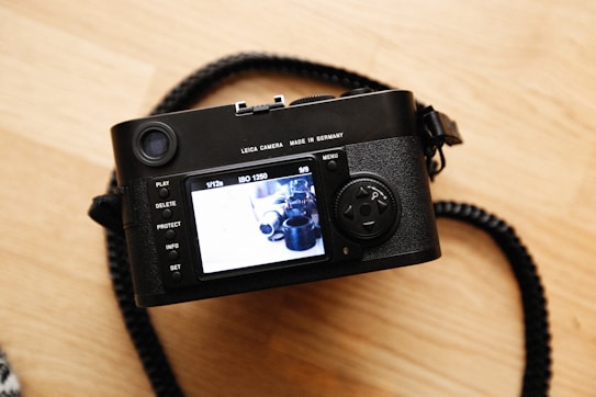 A digital camera is placed on a wooden surface, showcasing its display screen which is turned on. The camera is primarily black and has a strap attached to it. Various buttons, such as play, delete, and protect, are visible on the camera body next to the screen.