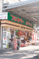 A small outdoor shop with a variety of items displayed, including umbrellas, bags, and hats. The storefront is vibrant with multiple red paper lanterns and bold signage in Japanese text. A vending machine stands to the left of the entrance.