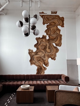 A modern interior space decorated with a large, abstract wooden wall art installation that has organic, swirling patterns. The seating area features a plush, brown sectional sofa with a textured surface, accompanied by two wooden coffee tables. A unique, spherical chandelier hangs from the ceiling, casting soft illumination. The room has a minimalist aesthetic with neutral tones and a sophisticated ambiance.