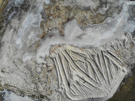 Aerial view of a large, barren landscape with distinct striations and layers, suggesting geological or mining activity. The scene is dominated by earthy tones and a textured surface resembling natural formations.