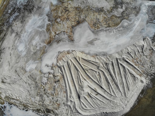 Aerial view of a large, barren landscape with distinct striations and layers, suggesting geological or mining activity. The scene is dominated by earthy tones and a textured surface resembling natural formations.
