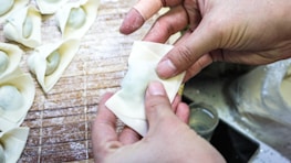 Hands are folding dumpling wrappers on a floured wooden board, creating neatly arranged dumplings. The surface is dusted with flour, and several partially folded dumplings are visible.