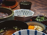A close-up view of a variety of Japanese dishes in different bowls and plates. There is a bowl of soup, chopped herbs, marinated yellow and white vegetables, and a hexagonal bowl with a blue pattern. The setting has a rustic wooden table and traditional Japanese dishware.