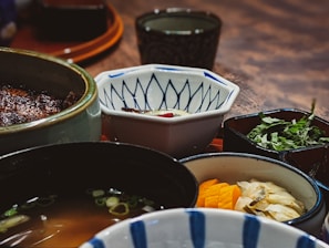 A close-up view of a variety of Japanese dishes in different bowls and plates. There is a bowl of soup, chopped herbs, marinated yellow and white vegetables, and a hexagonal bowl with a blue pattern. The setting has a rustic wooden table and traditional Japanese dishware.