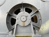 A close-up view of an industrial metal component featuring a circular vent or fan with several blades. The component is bolted onto a surface and has markings with numbers and letters on the metal. The surface appears aged with slight rust and wear.