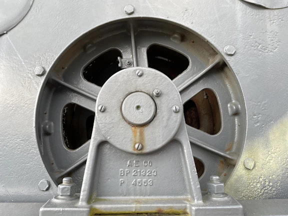 A close-up view of an industrial metal component featuring a circular vent or fan with several blades. The component is bolted onto a surface and has markings with numbers and letters on the metal. The surface appears aged with slight rust and wear.