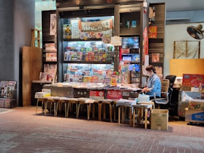 A small newsstand is filled with a variety of magazines, newspapers, and other reading materials. A person, seated on a chair, is handling something possibly related to the maintenance or organization of the kiosk. The newsstand is surrounded by stools and cardboard boxes, contributing to a cluttered yet lively street vendor atmosphere.