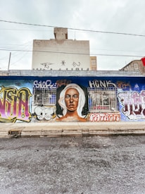 A mural painted on an urban street wall features a large portrait of a person with light hair. Surrounding the portrait are various vibrant graffiti styles and tags in different colors and fonts. The scene captures an urban environment with a building visible in the background and overhead wires.