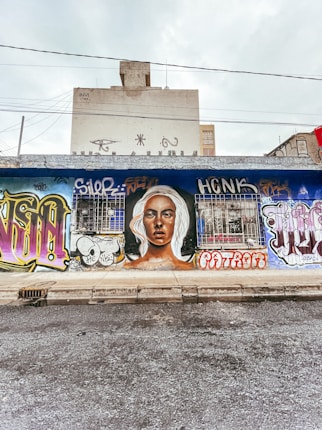 A mural painted on an urban street wall features a large portrait of a person with light hair. Surrounding the portrait are various vibrant graffiti styles and tags in different colors and fonts. The scene captures an urban environment with a building visible in the background and overhead wires.