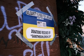 A poster on a wall, secured with duct tape, features the Ukrainian flag's blue and yellow colors and reads 'Solidarity is now for Ukraine'. It includes a call for donations to 'savethechildren.ch' and a QR code. The background has graffiti on a brown surface and there are plants with flowers on the right.