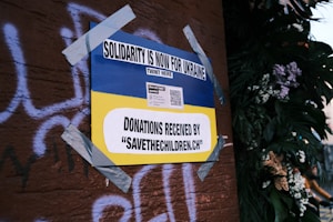 A poster on a wall, secured with duct tape, features the Ukrainian flag's blue and yellow colors and reads 'Solidarity is now for Ukraine'. It includes a call for donations to 'savethechildren.ch' and a QR code. The background has graffiti on a brown surface and there are plants with flowers on the right.