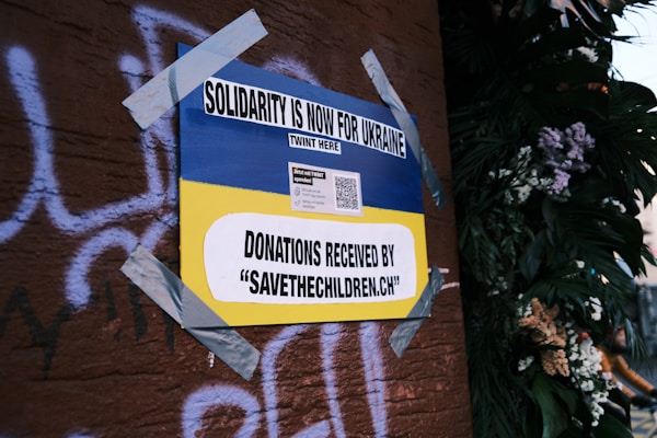 A poster on a wall, secured with duct tape, features the Ukrainian flag's blue and yellow colors and reads 'Solidarity is now for Ukraine'. It includes a call for donations to 'savethechildren.ch' and a QR code. The background has graffiti on a brown surface and there are plants with flowers on the right.