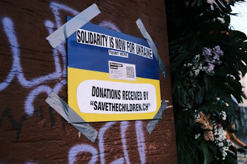 A poster on a wall, secured with duct tape, features the Ukrainian flag's blue and yellow colors and reads 'Solidarity is now for Ukraine'. It includes a call for donations to 'savethechildren.ch' and a QR code. The background has graffiti on a brown surface and there are plants with flowers on the right.
