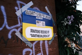 A poster on a wall, secured with duct tape, features the Ukrainian flag's blue and yellow colors and reads 'Solidarity is now for Ukraine'. It includes a call for donations to 'savethechildren.ch' and a QR code. The background has graffiti on a brown surface and there are plants with flowers on the right.