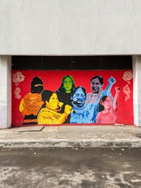 A vibrant mural painted on a red background, featuring multiple figures in various colors. The figures include a person with a covered face wearing a yellow and orange outfit, a person in green with glasses, and several others in blue, pink, and mixed tones. The artwork is accented with decorative white designs on the sides.