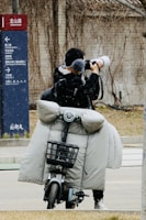 A person stands on a street taking photographs with a large camera. They are on a motorbike equipped with a grey, padded cover to protect against cold weather. A directional sign appears on the left side indicating different places.