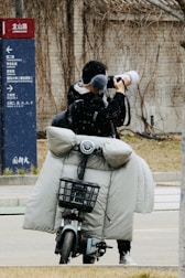 A person stands on a street taking photographs with a large camera. They are on a motorbike equipped with a grey, padded cover to protect against cold weather. A directional sign appears on the left side indicating different places.