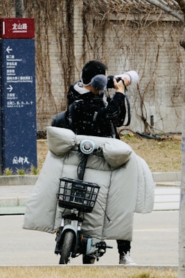 A person stands on a street taking photographs with a large camera. They are on a motorbike equipped with a grey, padded cover to protect against cold weather. A directional sign appears on the left side indicating different places.