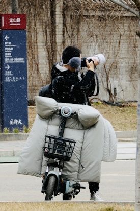 A person stands on a street taking photographs with a large camera. They are on a motorbike equipped with a grey, padded cover to protect against cold weather. A directional sign appears on the left side indicating different places.