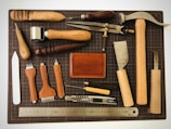 An assortment of leatherworking tools neatly arranged on a cutting mat. The collection includes tools such as hammers, awls, chisels, a cutting knife, and a steel ruler. A mid-sized leather wallet lies in the center amidst the tools, and various leather punches with wooden and metal handles are also present. The items are organized to display a variety of materials used in leather crafting.