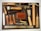 An assortment of leatherworking tools neatly arranged on a cutting mat. The collection includes tools such as hammers, awls, chisels, a cutting knife, and a steel ruler. A mid-sized leather wallet lies in the center amidst the tools, and various leather punches with wooden and metal handles are also present. The items are organized to display a variety of materials used in leather crafting.