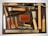 An assortment of leatherworking tools neatly arranged on a cutting mat. The collection includes tools such as hammers, awls, chisels, a cutting knife, and a steel ruler. A mid-sized leather wallet lies in the center amidst the tools, and various leather punches with wooden and metal handles are also present. The items are organized to display a variety of materials used in leather crafting.