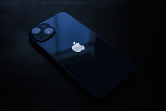 A sleek, dark-colored smartphone lying flat on a dark surface. The device features a prominent logo on the back, two visible camera lenses, and reflects a soft ambient light.