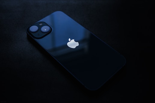A sleek, dark-colored smartphone lying flat on a dark surface. The device features a prominent logo on the back, two visible camera lenses, and reflects a soft ambient light.