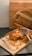 A partially eaten baked dish in a glass casserole dish sits on a wooden board, accompanied by a metal spatula and fork. The setting includes a round wooden cutting board tilted against a wooden backdrop, creating a warm and rustic kitchen scene.