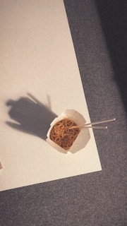 A takeout container filled with noodles rests on a surface, accompanied by chopsticks. The shadow of a hand is visible nearby, cast on the surface.