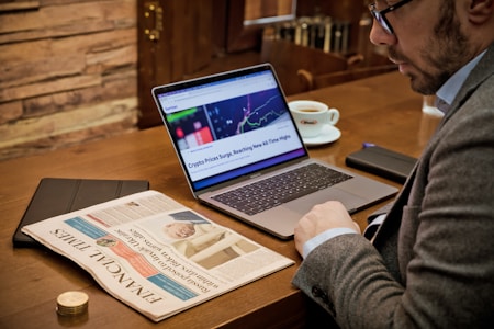A man sits at a table in a cafe or office environment, wearing glasses and a suit. In front of him, an open laptop displays a webpage about cryptocurrency price surges. A newspaper titled 'Financial Times' lies on the table, along with a cup of coffee and a smartphone. The table surface is wooden, contributing to a cozy, businesslike atmosphere.