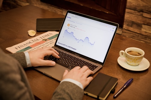 A person is working on a laptop displaying a cryptocurrency price chart. A newspaper is placed on the wooden table next to a cup of coffee, a notebook, and a pen.