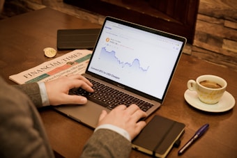 A person is working on a laptop displaying a cryptocurrency price chart. A newspaper is placed on the wooden table next to a cup of coffee, a notebook, and a pen.
