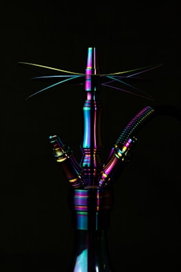 A metallic hookah with a rainbow-like iridescent finish is prominently displayed against a dark background. The lighting accentuates the reflective surface, creating a spectrum of colors including purple, green, and blue. The sleek design includes multiple ports and a ribbed hose attached.