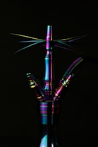 A metallic hookah with a rainbow-like iridescent finish is prominently displayed against a dark background. The lighting accentuates the reflective surface, creating a spectrum of colors including purple, green, and blue. The sleek design includes multiple ports and a ribbed hose attached.