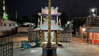 A nighttime scene of a brightly lit, ornate temple with intricate carvings and a golden pole in the foreground. There are colorful decorations and religious statues present, surrounded by a paved courtyard and some plastic containers on the ground.