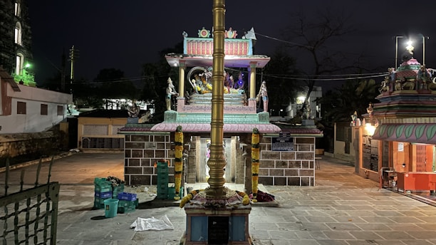 A nighttime scene of a brightly lit, ornate temple with intricate carvings and a golden pole in the foreground. There are colorful decorations and religious statues present, surrounded by a paved courtyard and some plastic containers on the ground.