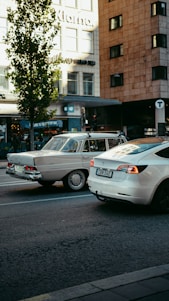 A modern white Tesla car is parked next to a vintage beige car on a city street. The background features urban buildings with large windows and signage, and a tree is positioned near the vehicles, casting a shadow on the asphalt. The scene suggests a juxtaposition between modern and classic automotive designs.