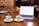 A wooden table holds a laptop displaying a cryptocurrency price chart, two cups of coffee, a smartphone, and a magazine with coins on top. A person is interacting with a smartphone on the right side.