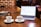 A wooden table holds a laptop displaying a cryptocurrency price chart, two cups of coffee, a smartphone, and a magazine with coins on top. A person is interacting with a smartphone on the right side.