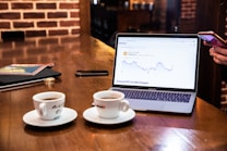 A wooden table holds a laptop displaying a cryptocurrency price chart, two cups of coffee, a smartphone, and a magazine with coins on top. A person is interacting with a smartphone on the right side.