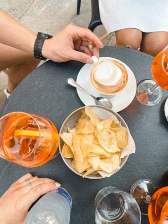 A casual dining setup is portrayed with a dark round tabletop, featuring a cappuccino cup and saucer, alongside a spoon placed nearby. A bowl filled with crispy potato chips is centrally positioned. Around the table are two bright orange-hued beverages with slices of lemon and a hand holding a bottle of sparkling water. Partial views of individuals sitting give a relaxed and social ambiance.