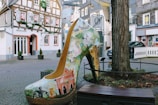 A large, decorative high-heeled shoe sculpture is situated in a quaint European street setting. The shoe is adorned with vibrant murals depicting architectural landmarks and natural landscapes. The background features charming half-timbered buildings with festive decorations, cobblestone pavement, and a large tree. Various signs and a parked car can be seen in the vicinity, enhancing the urban environment.