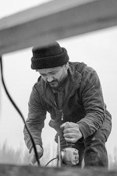 A black and white photo of a bearded man wearing a beanie and working with a power tool. He appears focused, leaning over the tool which is on the ground, and dust covers his clothing. The environment is outdoors, and the blurred background suggests a possibly remote or industrial setting.