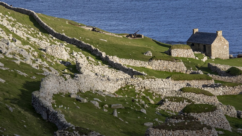 an old stone house on a grassy hill next to a body of water