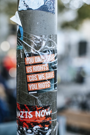 A pole is covered with multiple layered stickers. One sticker reads, 'YOU ONLY GAVE US RIGHTS 'COS WE GAVE YOU RIOTS' in bold orange and white text. Another sticker below reads 'NAZIS NOW' in white, set against a red and black background with graphic illustrations. A sticker at the top mentions 'Fridays for Future,' featuring a globe and a hand graphic.